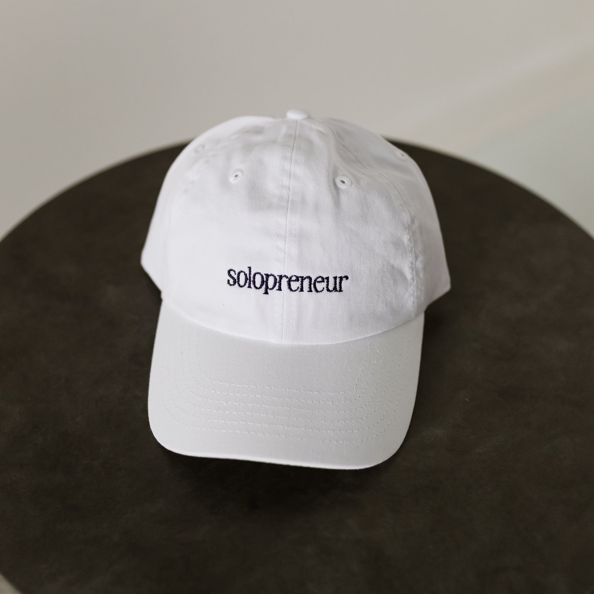 white baseball cap embroidered with navy thread "Solopreneur" on a table