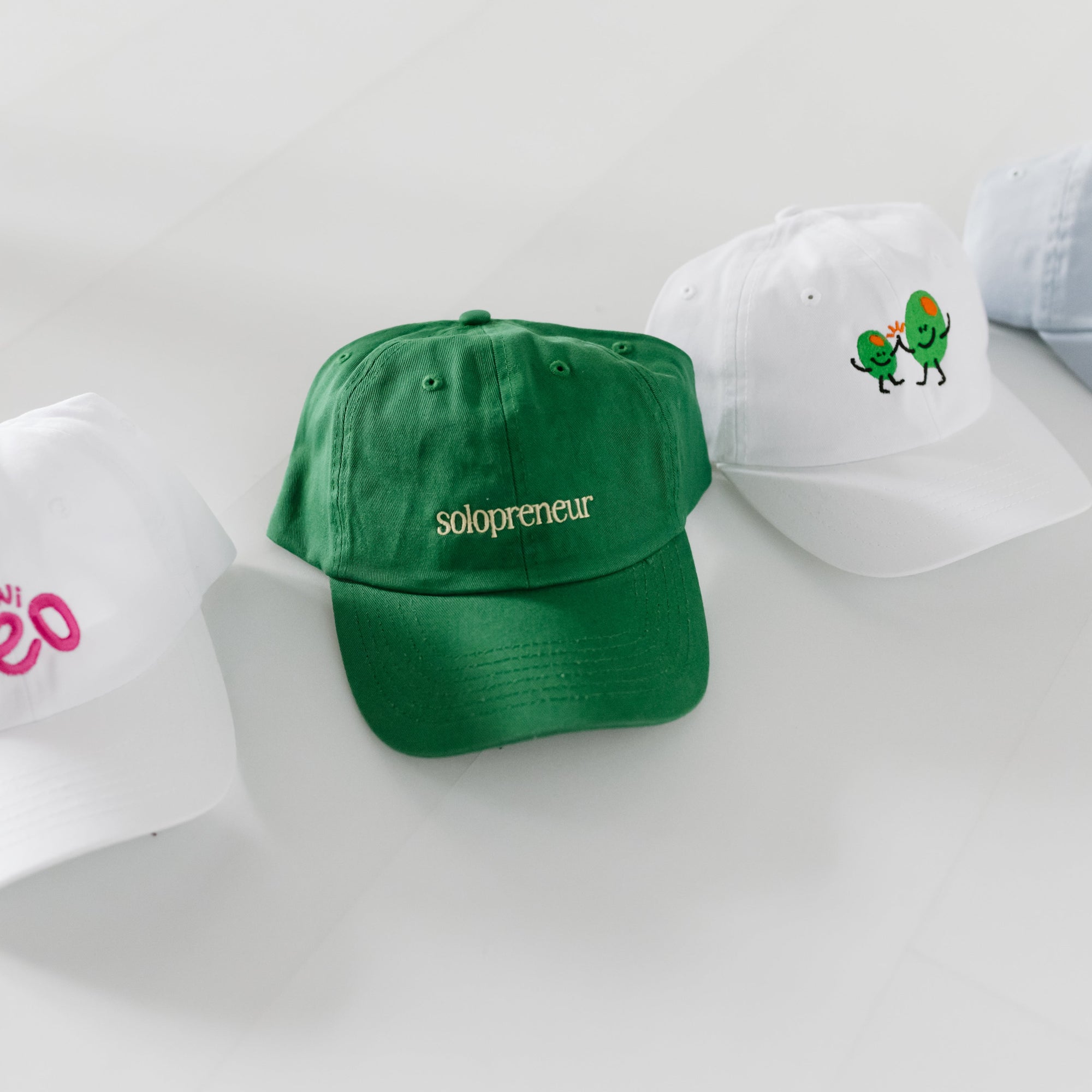 green baseball cap embroidered with solopreneur text next to other fun baseball caps for toddlers and kids